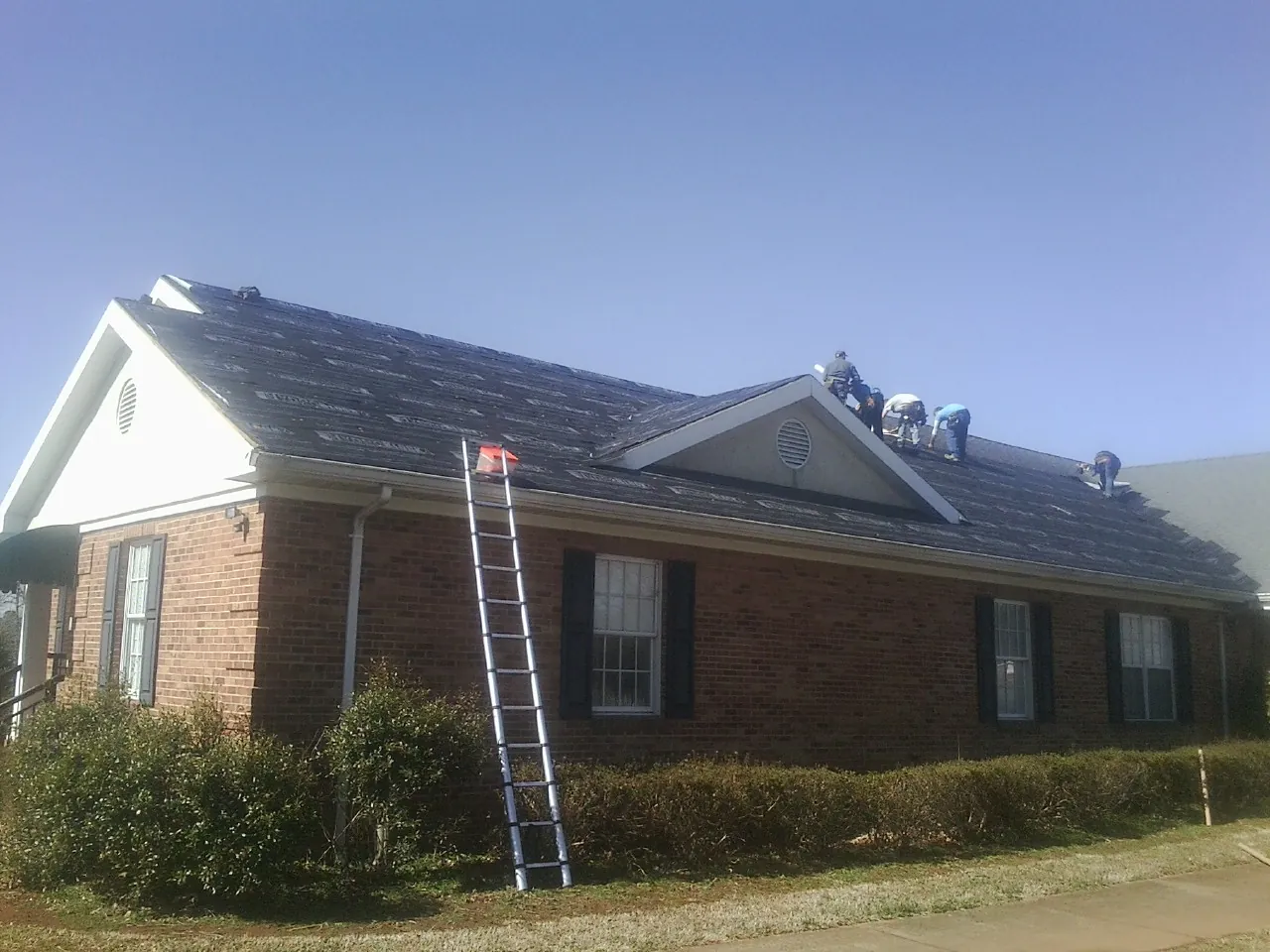 Crew tearing shingles off a brick ranch roof with a ladder set — Metal Roof Repair work by our crew in Churchville